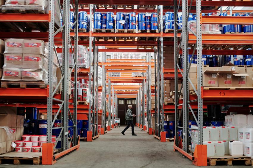 An image of filled shelves in a warehouse, taken from the angle of down one of the aisles, with a person walking through an adjacent aisle.