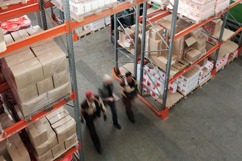 An aerial image of three people in safety uniforms and hats walking down an aisle in a warehouse, with stocked shelves on either side.