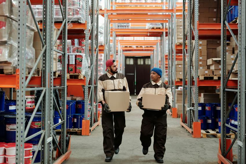 An image of a warehouse with filled shelves, taken down the length of one aisle. There are two male warehouse workers walking towards the camera, each carrying a box.