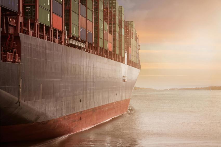 A large barge, filled with green, blue and red shipping containers, sailing on the sea towards a sunset.