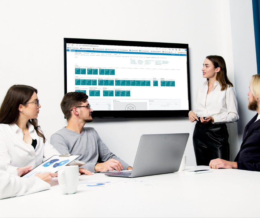 Three people in a meeting room in an office, two men and two women. One woman is standing next to a wall-mounded screen showing a Dynamics 365 Business Central dashboard.