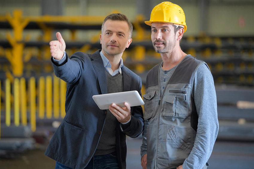 Two men in a manufacturing warehouse. One is wearing a suit and gesturing forward, holding a tablet. The other is wearing a hard hat and overalls.