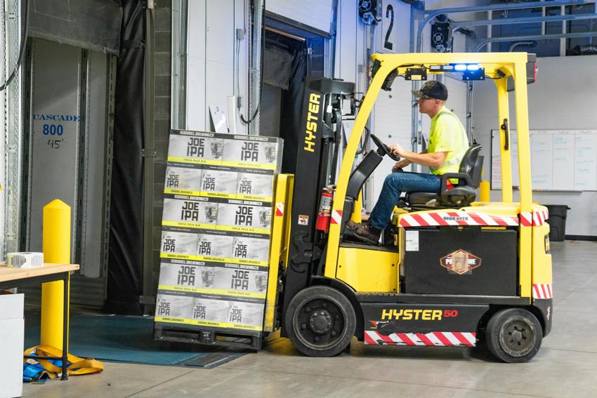 A man controlling a yellow forklift truck with a pallet of items on it in a warehouse.